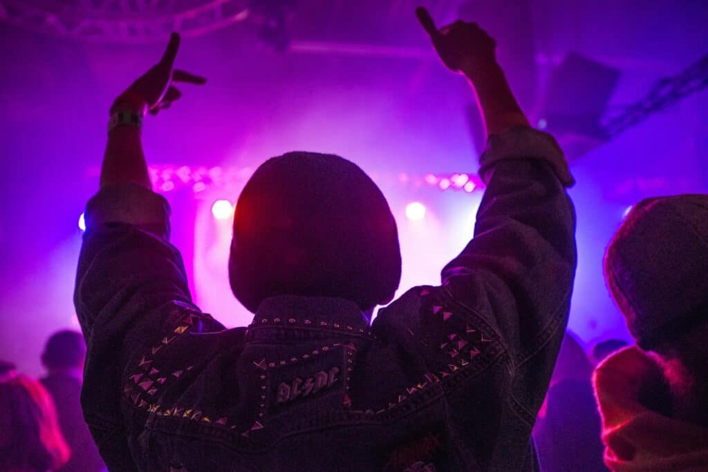 back of man raising hands inside room full of people with purple lights